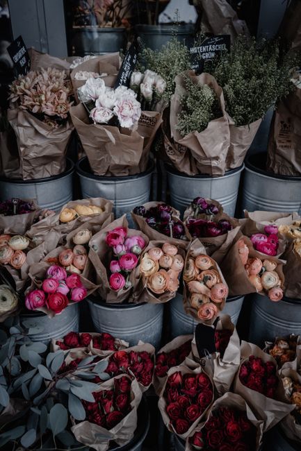 A display of fresh flowers arranged in metal buckets and paper-wrapped bouquets at a flower shop, showcasing a variety of roses in shades of pink, peach, and red, along with delicate white blooms and