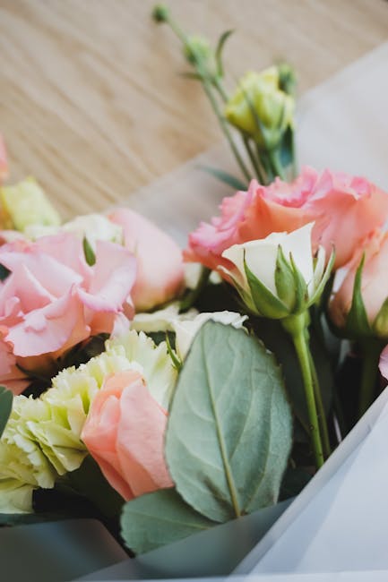 Close-up of a floral bouquet featuring soft pink and cream Lisianthus flowers with layered, ruffled petals, complemented by green leaves and stems. The bouquet is partially wrapped in light-colored, s