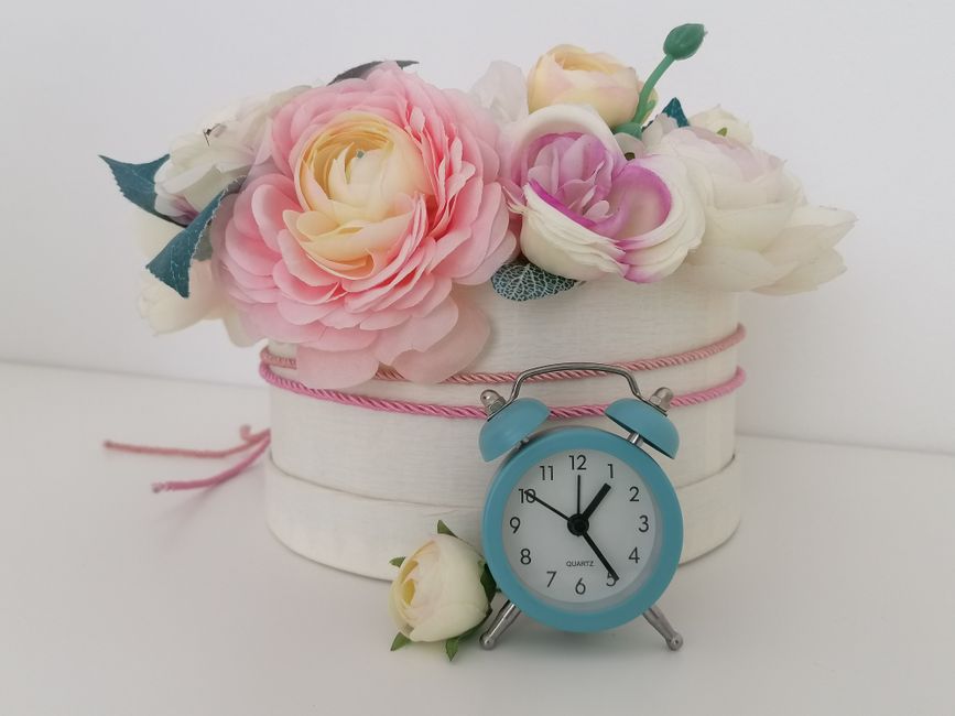A floral arrangement featuring a bouquet of artificial roses in soft pink, cream, and lavender hues, with textured petals and green leaves, placed inside a white, oval-shaped box wrapped with pink and