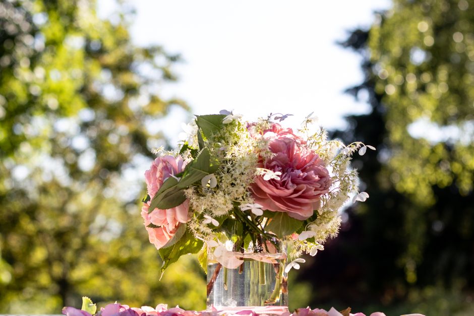 A floral arrangement of large, soft pink peonies with ruffled petals, accented by small white filler flowers and lush green leaves, all arranged in a clear glass vase. The bouquet is placed outdoors o