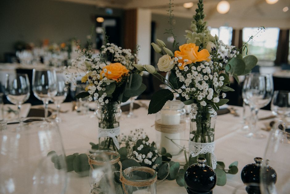 A floral arrangement featuring a bouquet of fresh flowers displayed in glass jars on a table set for an event. The bouquet includes vibrant yellow roses, white roses, and delicate white baby's breath,